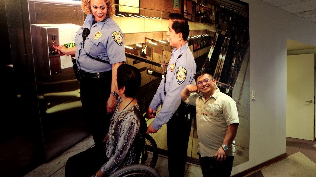 A man smiles in front of a picture of a three people with one of them in a wheelchair in front of an elevator.