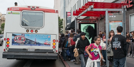 Rear of Muni bus stopped at bus stop with a large number of people crowding towards the rear doors and waiting at the stop and on the sidewalk