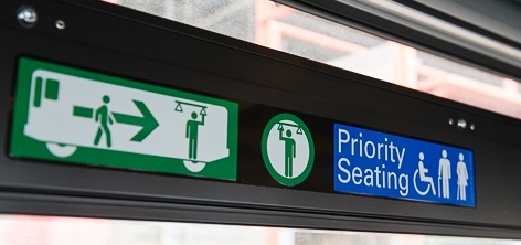 detailed color photograph showing graphic decals on Muni bus. the decals, which are blue and white and green and white show stylized figures moving towards rear of stylized bus and have the words "Priority Seating" showing a wheelchair symbol.