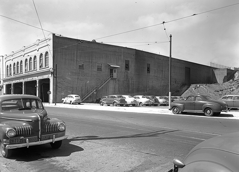 black and white photo from 1948 showing the rear side of Muni's Presidio Division. The rear wall of the building has one angled corner instead of being a perfect rectangular shape. Behind the building is a partially excavated hillside, the former location of Laurel Hill Cemetery.