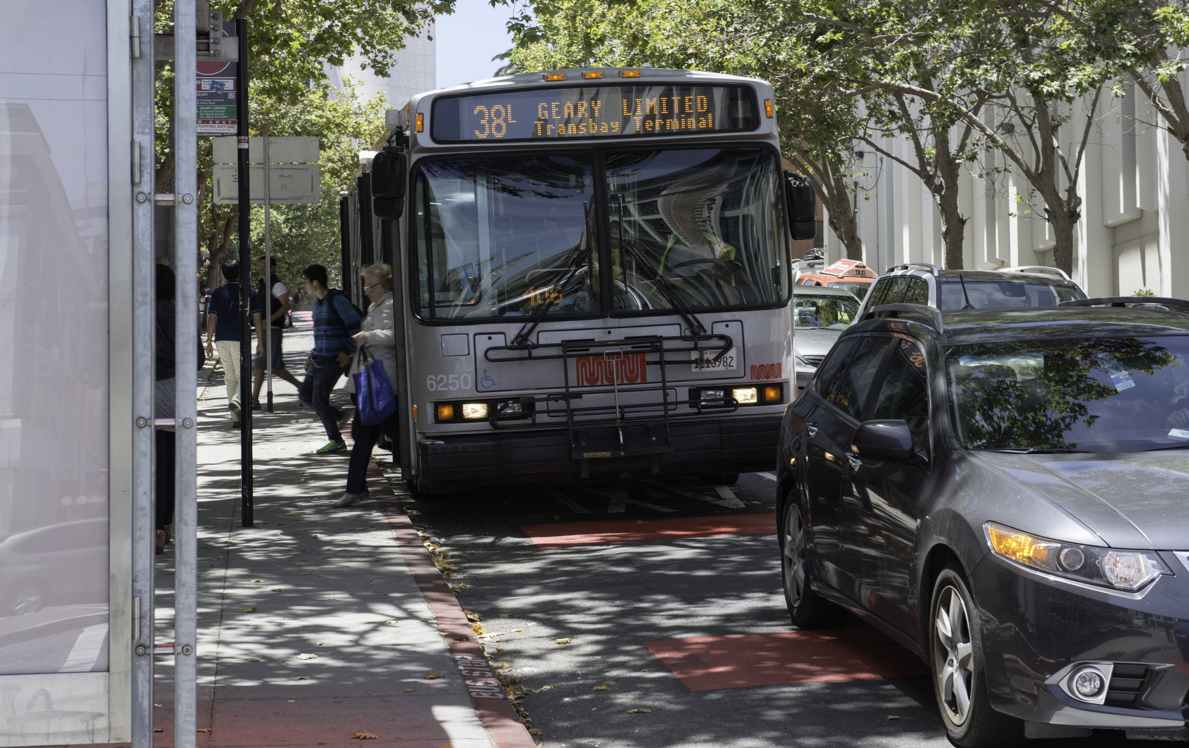 Front of Muni 38L Geary Limited bus at curb, off-loading customers on a downtown street with car traffic around it.