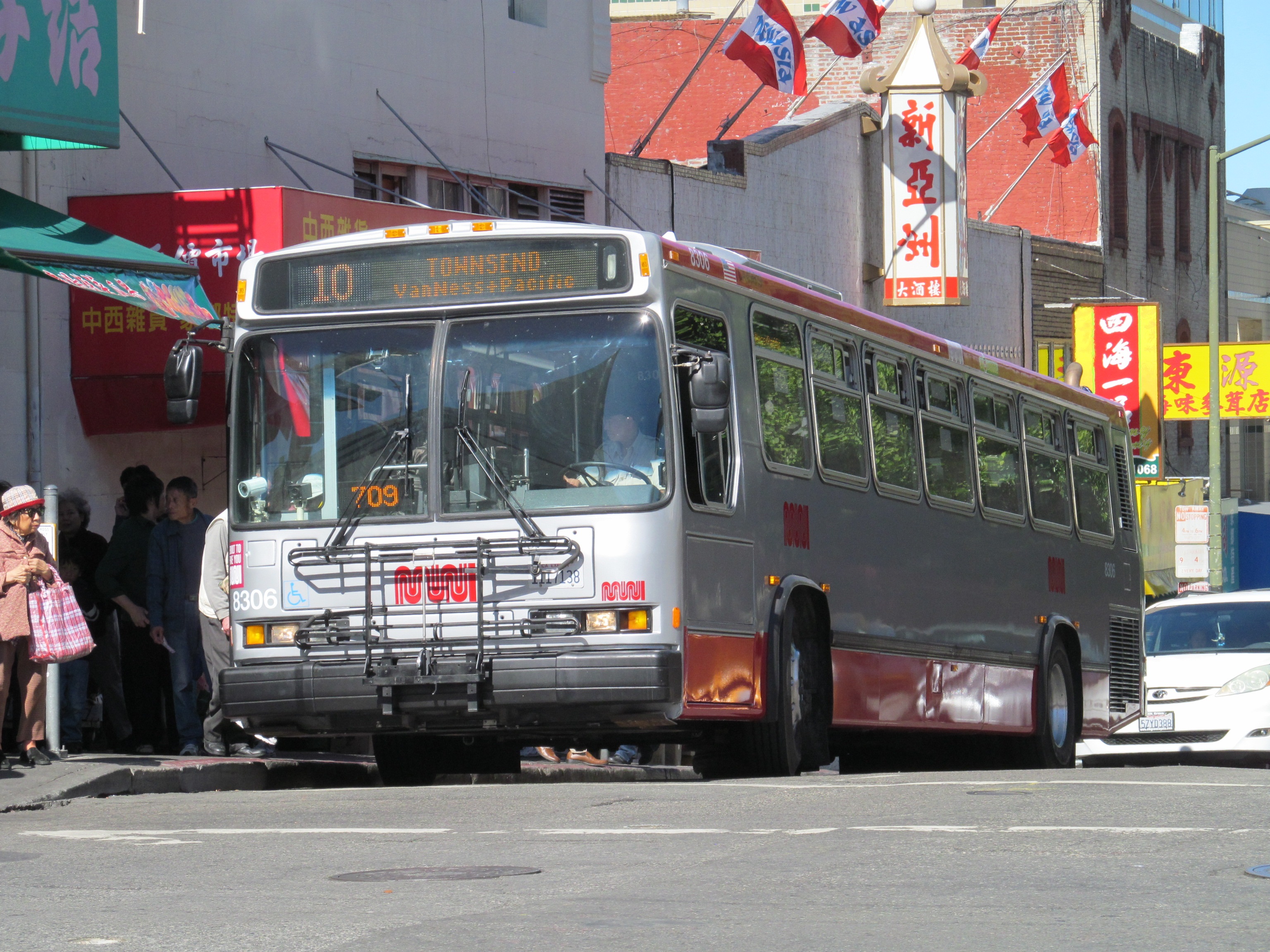 10 Townsend silver and red Muni bus waits at the curb for passengers to board with colorful neighborhood banners and signs in the background.
