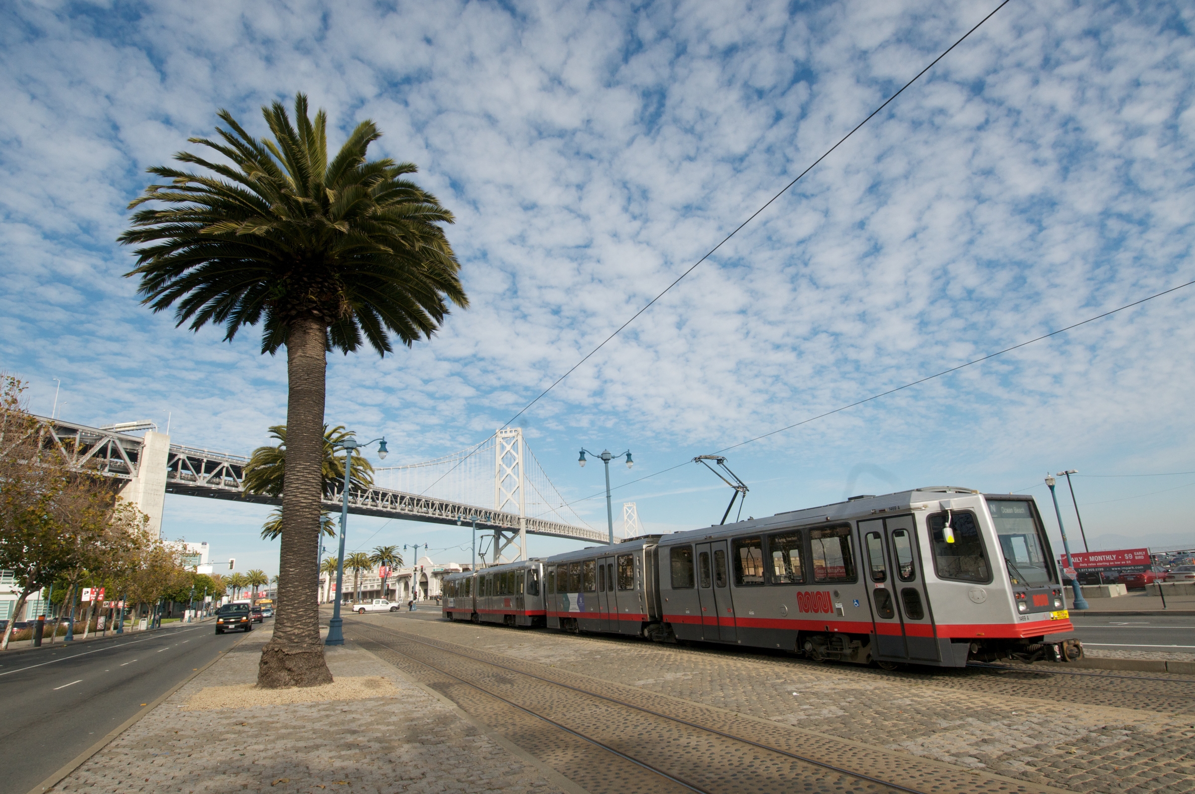 An N Judah light rail train travels north on The Embarcadero with a palm tree on the left and the Bay Bridge in the background.