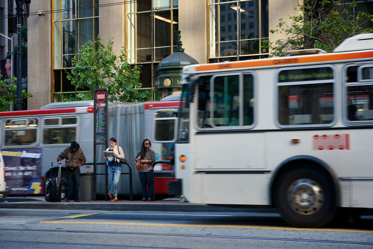 Muni buses pass on Market Street as one pulls up to waiting passengers on boarding island.