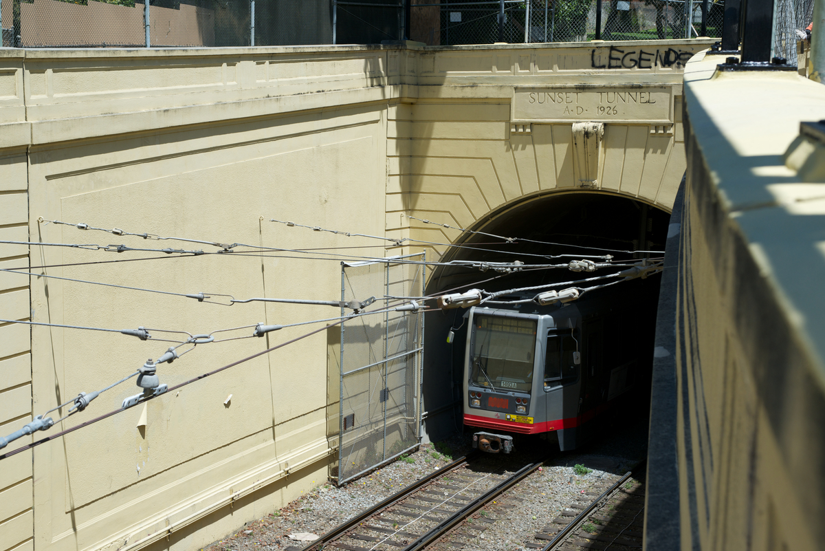 Gray and red light rail train exits westbound from the sunken Sunset Tunnel portal into Cole Valley.