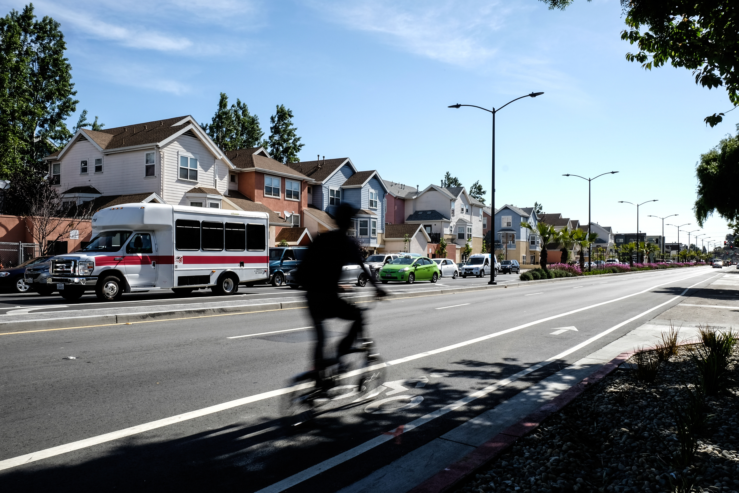 A bicyclist in shadow rides along the new Cesar Chavez street with traffic and houses in the background.