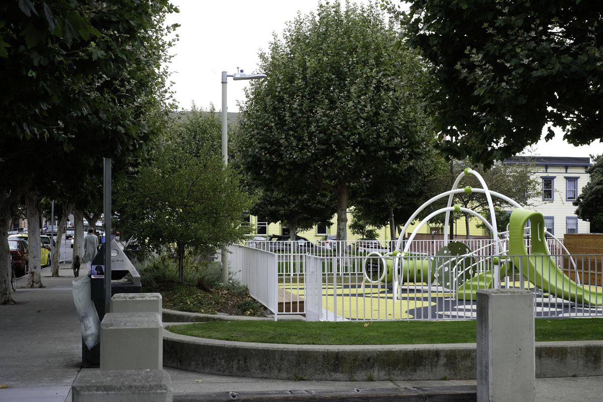 Children's green and white play equipment in a fenced-in playground on a tree-lined street.