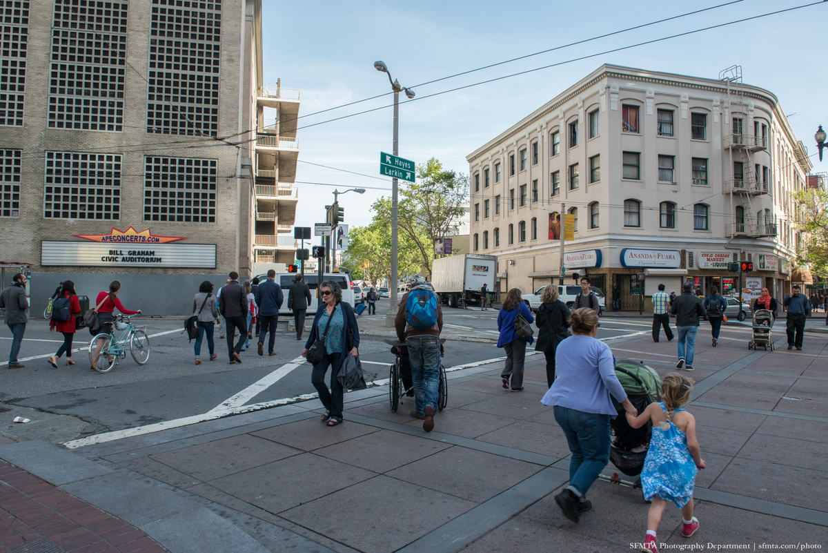 Pedestrians with strollers, a wheelchair and a bicycle all traverse the intersection of Market, Hayes and Larkin streets