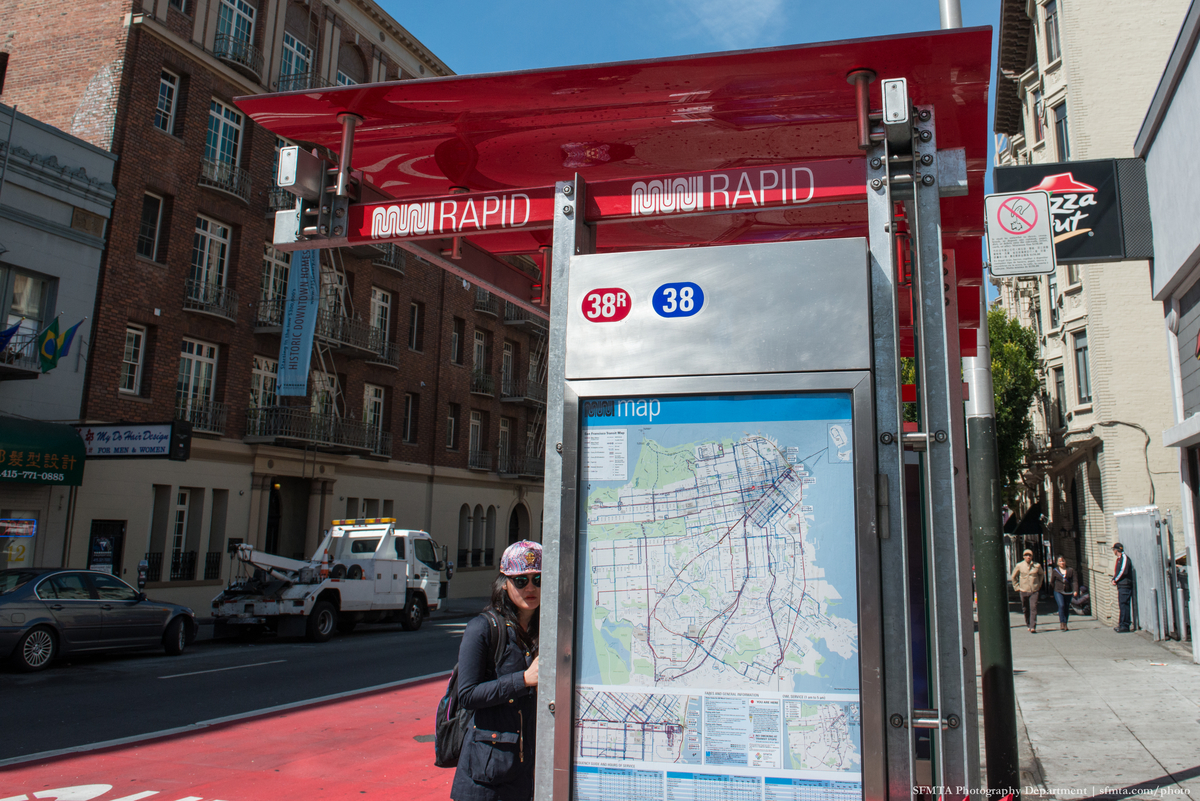 Woman stands next to Muni shelter with red "Muni Rapid" decals and new map with red and blue route lines.