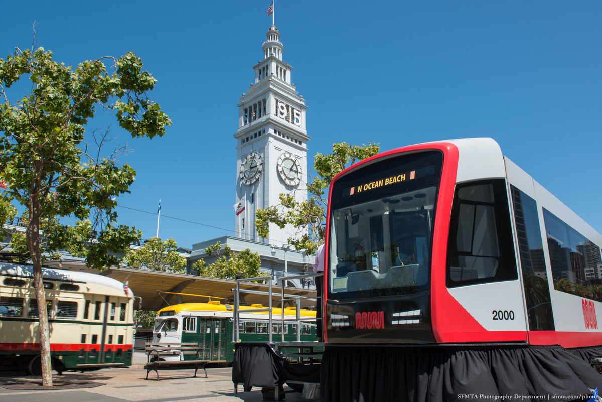Replica gray and red light rail train model sits on boarding island on The Embarcadero with a blue sky, the Ferry Building and colorful historic streetcars in the background.