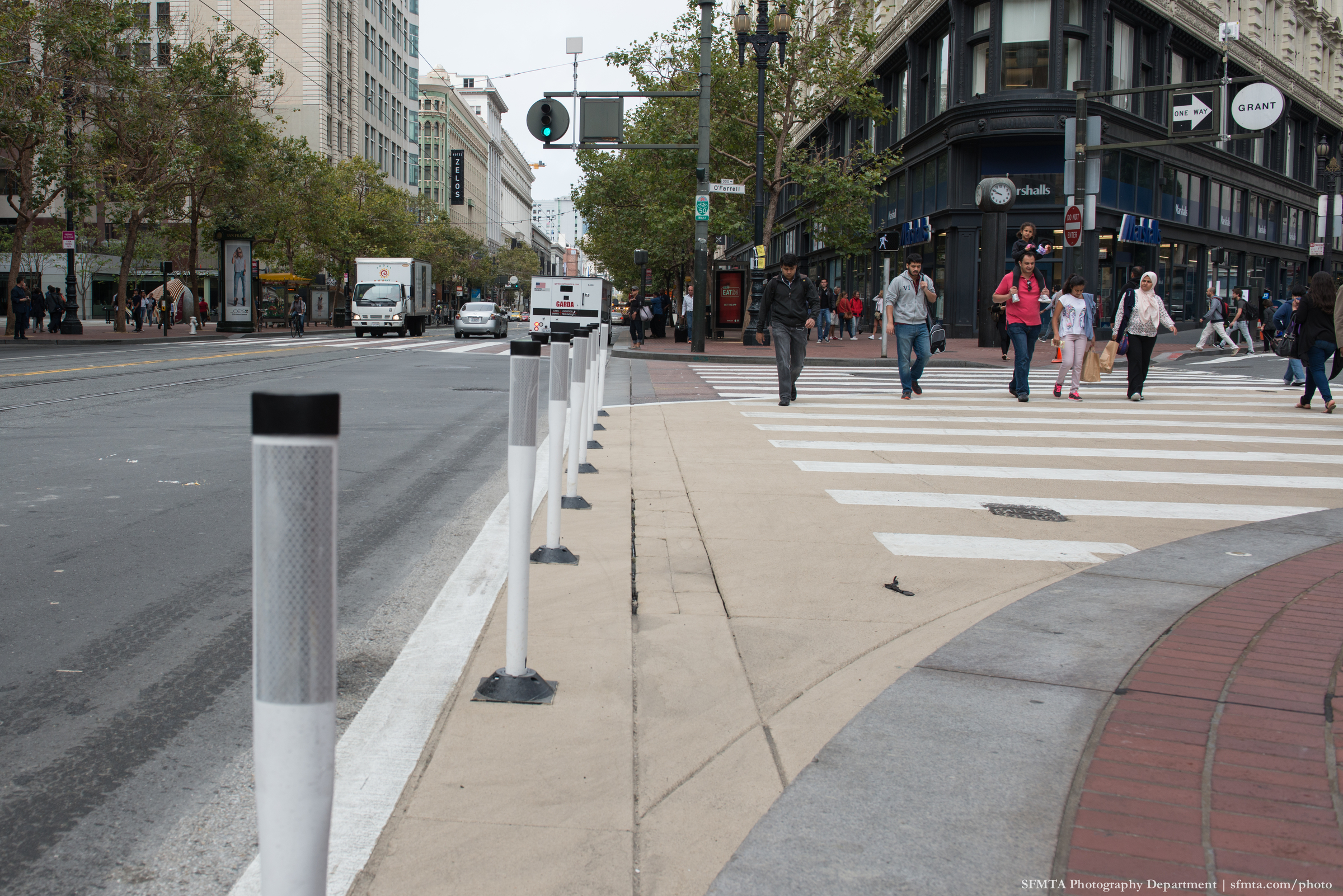 Pedestrians walk thorugh a zebra crosswalk at O'Farrell and Market where a newly installed buffer zone of beige paint and white plastic posts has been installed.