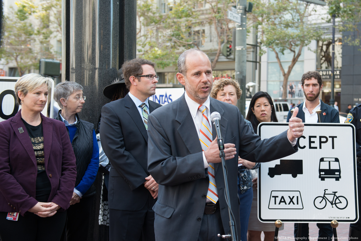 Ed Reiskin stands at microphone on Market at O'Farrell with turn-restriction signs and dignitaries behind him.