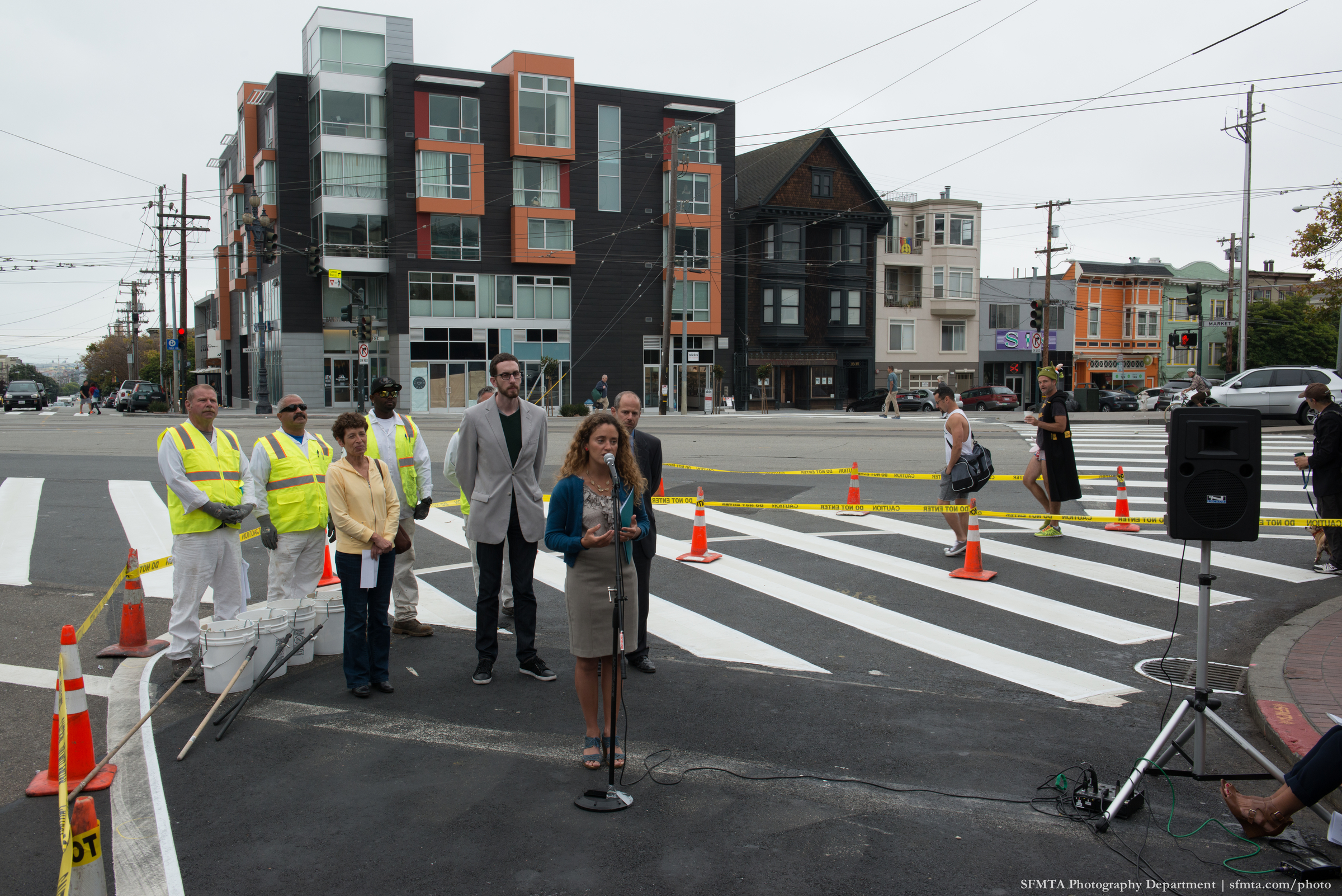 Dignitaries are gathered behind a microphone next to a crosswalk for a safety press conference, while a man in a batman t-shirt, a black knee-lenth vest, a green kid's knit hat, tennis shoes, and no pants walks through the crosswalk.