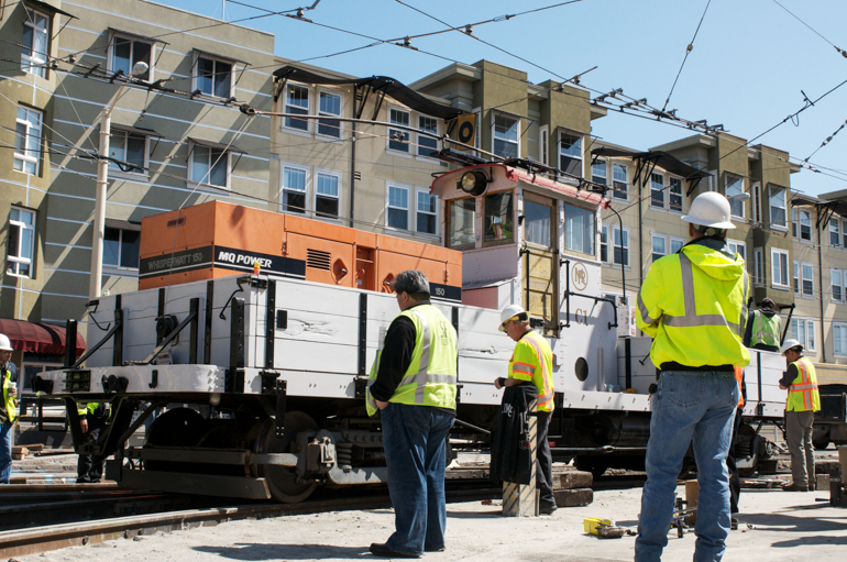 Work Car C-1 running over new rail junctions with workers standing nearby, street is unpaved.  Taken May 30, 2012 at Church Street and Duboce Avenue