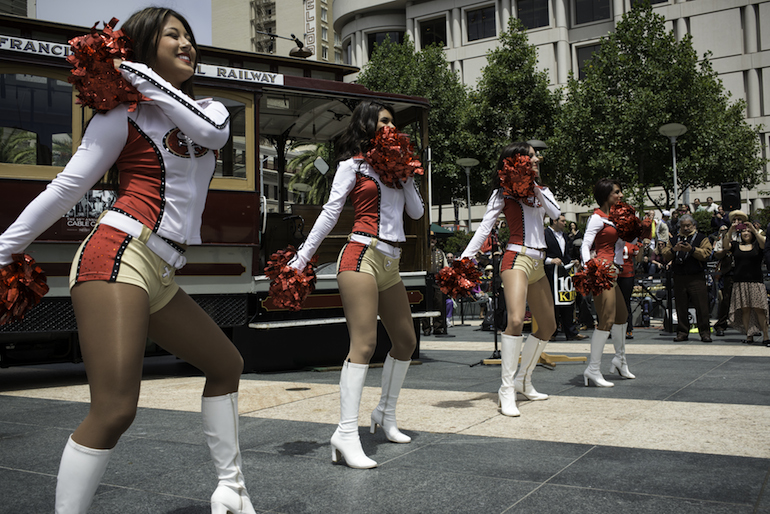 Group of athletic women in cheerleader costumes dancing in a row in front of Motorized Cable Car 62 in Union Square