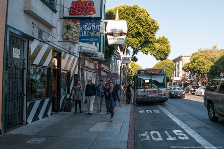 A white Muni bus sits at the curb on a crowded city street with pedestrians strolling past and colorful signs above the stores.