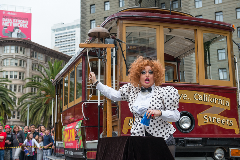 Color photograph of a woman wearing a black and white polka dot dress ringing a cable car bell while performing a magic trick involving blue cups and red balls at the 52nd annual bell ringing contest on July 9, 2015.