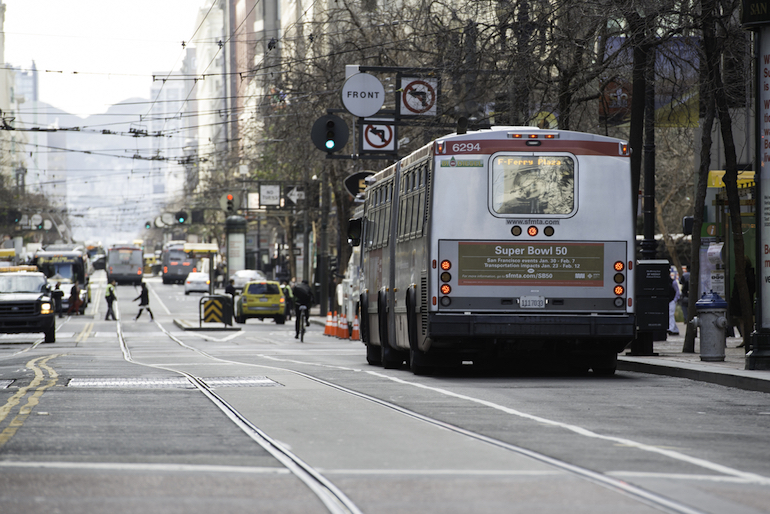 Muni bus with an SFMTA gold "Super Bowl 50" information PSA on the back travels down Market Street with pedestrians and other traffic around it.