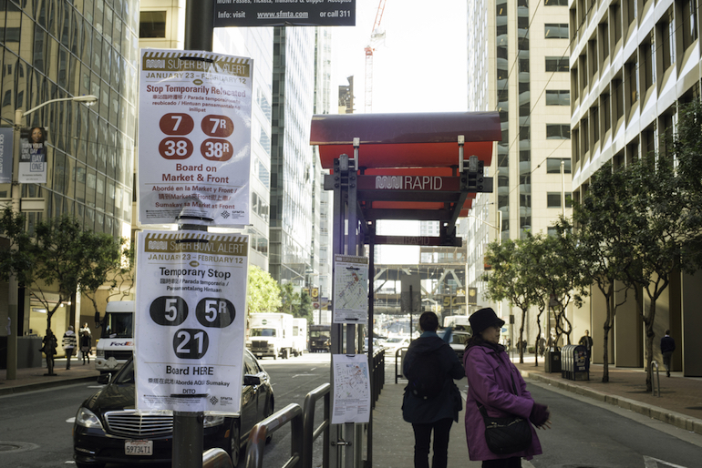 Red-topped Muni bus shelter on a busy downtown street with customers waiting and laminated "Super Bowl" alerts posted on it.