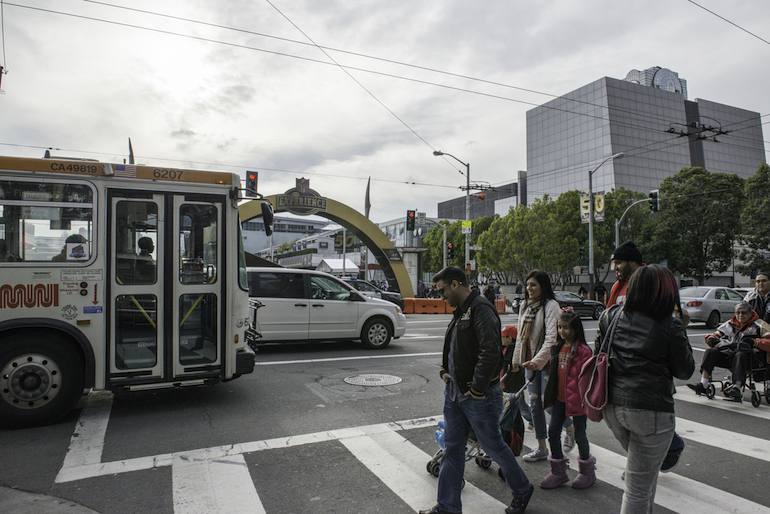 A Muni bus travels north on 3rd Street in front of the Super Bowl 50 NFL Experience entrance on Howard Street. Pedestrians cross southbound.