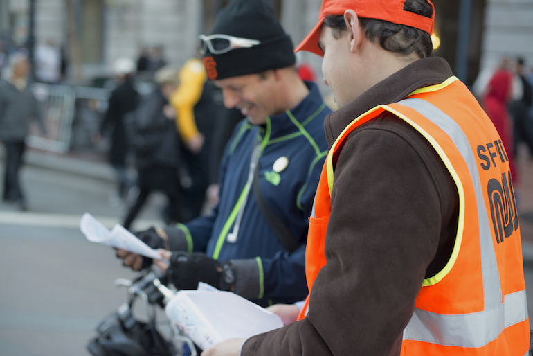 A Muni ambassador in safety orange vest and cap directs a visitor to SF during Super Bowl City.