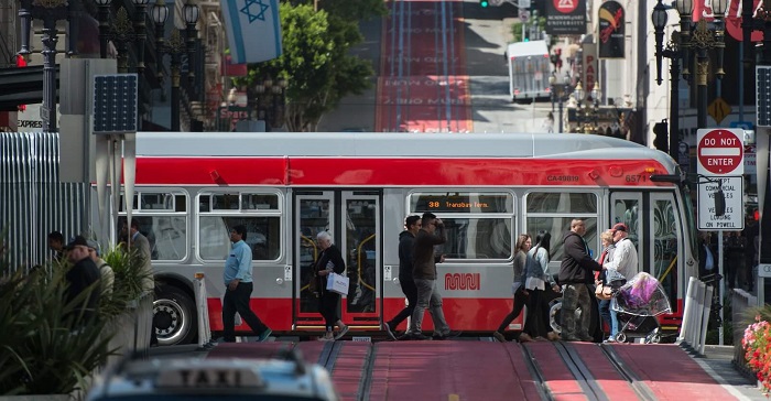 Gray and red articulated Muni bus traveling east on O'Farrell with the red transit lanes for cable cars in the background and foreground. Pedestrians cross in front of the bus.