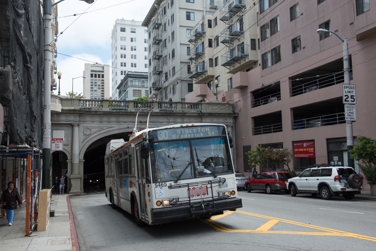 Color photo of an electric trolley bus exiting the northern portal of the Stockton Tunnel on Muni's 30 Stockton route at Stockton and Sacramento streets. The tunnel portal is arched with decorative smooth pillars on either side and a colonnade at top. Taken June 6, 2016.