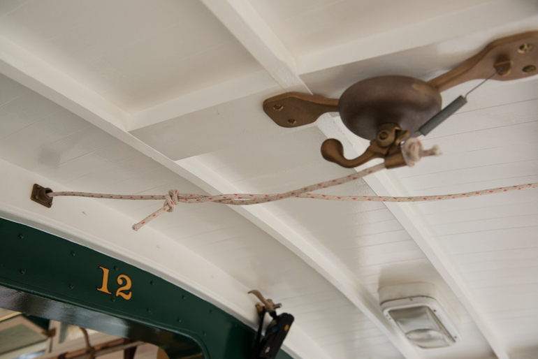 Color detail view of a cable car conductor's bell. This is a small brass bell mounted to the ceiling joist of the rear platform of a cable car. The bell has a rope attached to it which runs the length of the car and is used by the conductor to communicate with the grip person located at the front of the car. Also visible is the bulkhead of the car above the rear doorway with the gold-painted car number "12" painted on a green background. Photo taken June 16, 2016.