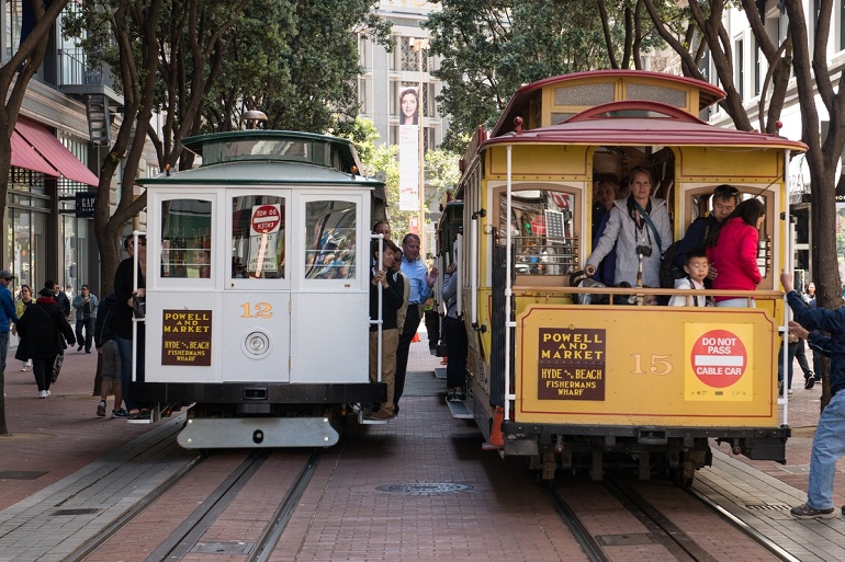 The white Cable Car 12 sits next to the gold Cable Car 15 on the tree-lined tracks of Powell Street, just south of Ellis.