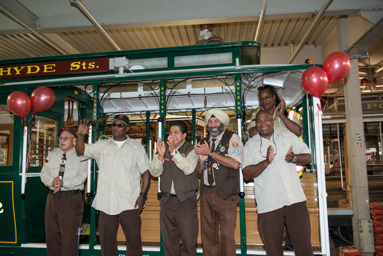 Group photo of seven Cable Car Bell Ringing Contest standing on running boards and side of cable car, clapping and smiling.