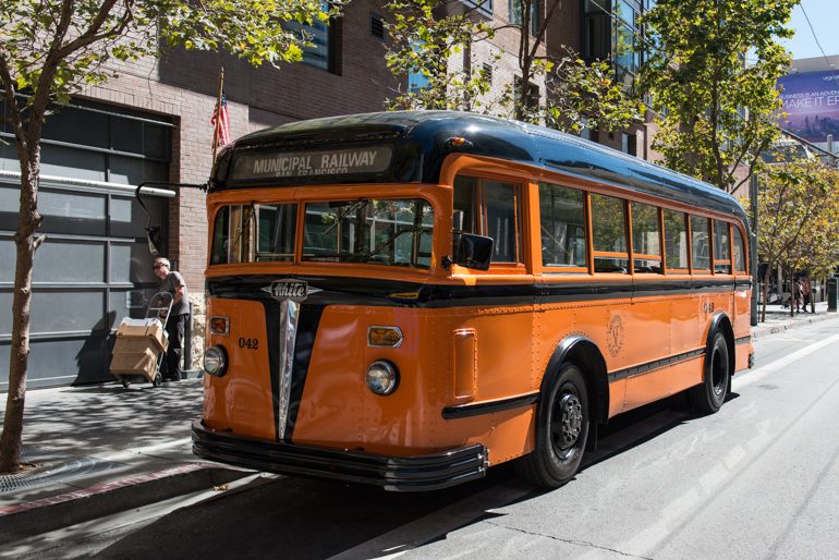 3/4 angle front view of 1938 Muni bus numbered 042.  This is a short bus painted in stark black and orange and features a strong vertical, spear-shaped chrome trim down the center of the front, beneath the split windscreen.