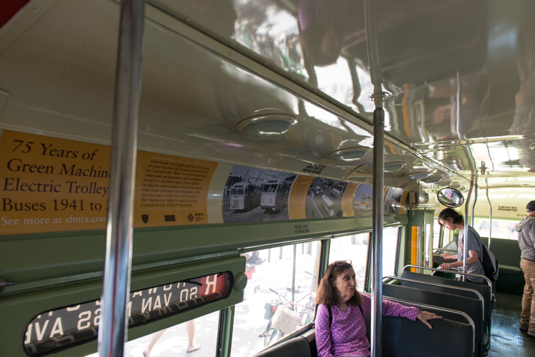 Interior view inside trolley coach 506 showing photo cards depicting the 75 year history of Muni trolley coaches. The bus has a light green ceiling and dark green vinyl seating set perpendicular to the walls of the bus. A woman wearing a purple sweater is seated in the lower right corner of the frame.