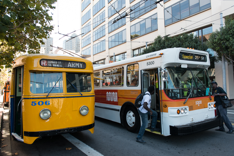Color photograph showing two electric trolley buses on Steuart Street.  One bus in the left foreground is painted a bright blue and yellow and wears the number "506". 506 is being passed by another 1970s bus, #5300 painted white, orange, and red.  A passenger wearing a white t-shirt boards #5300.