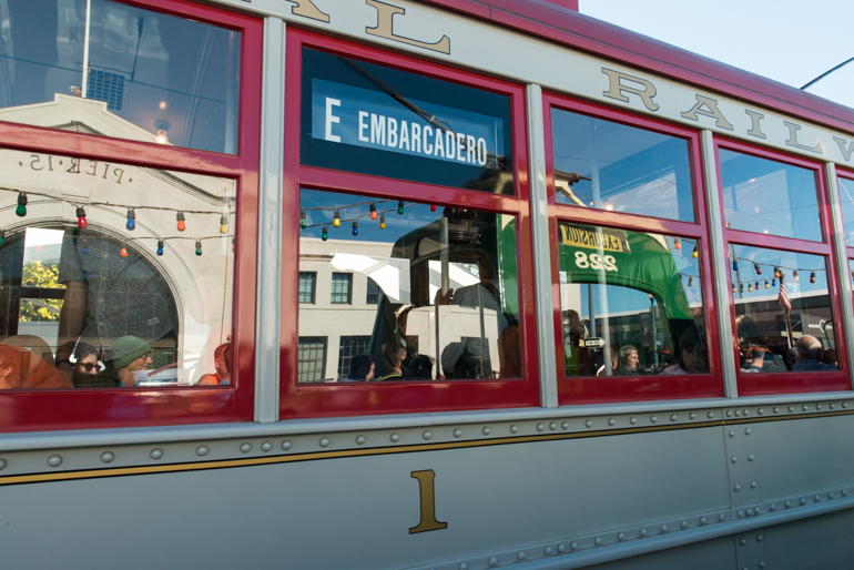 Side view of Muni's Streetcar 1, showing windows and red and grey paint job.  In the windows, a reflection of the open-air streetcar 228 can be seen along with a reflection of the facade of Pier 15 on the Embarcadero.