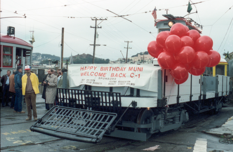 Color photograph of Work streetcar C-1 on Market Street and Duboce Avenue.  Car is a flatbed streetcar with a control cabin in the center.  Restored for Muni's 80th birthday.  Taken December 28, 1992
