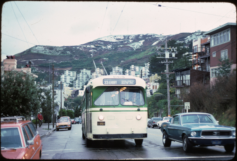 a color photograph from February 1976 showing a Muni electric trolley coach operating on the 33 Ashbury line on Clayton Street near Deming Street.  the bus and a 1960s model Ford Mustang car are in the mid ground and Twin Peaks, dusted with white snow is seen in the background.