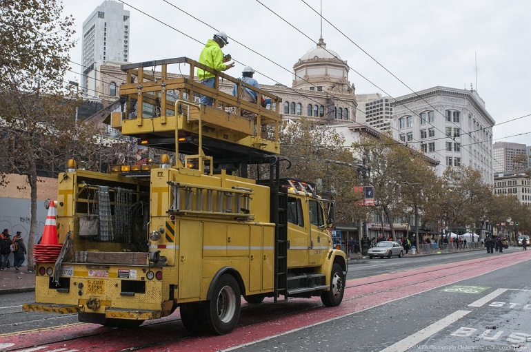 Workers stand on yellow truck's elevated platform to reach overhead catenary wires on Market Street. 