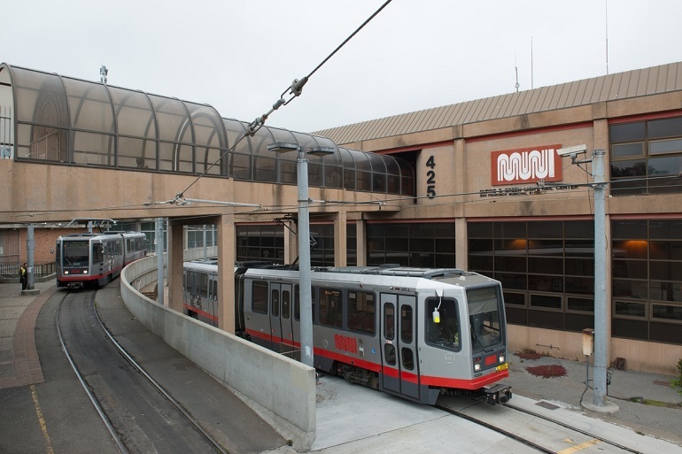 Large rail yard with enclosed walkway on the left and two light rail vehicles crossing below.
