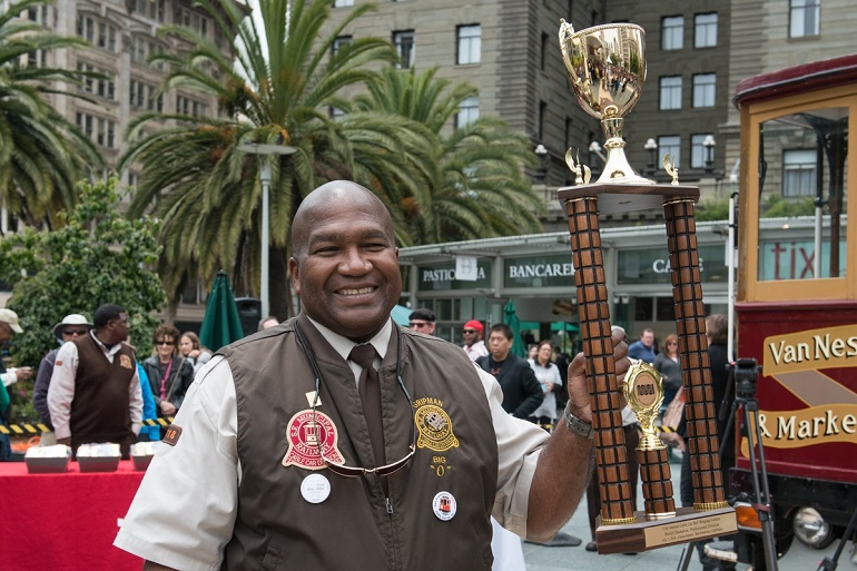 Muni Cable Car Operator Leonard Oats holds up his first place trophy in Union Square with the motorized cable car and palm trees in the background.