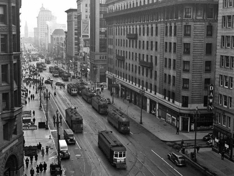 A 1940 photo shows a view above Market Street, looking east from Kearny Street, with crowded rows of streetcars running on four sets of tracks, along with pedestrians and automobiles. In the distance, the Ferry Building is just visible through the haze