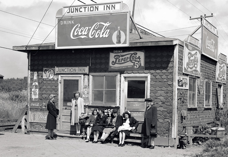 A black and white photograph from May 1945 shows a group of women sitting and standing in front a small building with a sign that reads “Junction Inn.” The building is covered with advertisements for Coca Cola and Pepsi Cola. The women are dressed in 1940s fashion with headscarves, long overcoats and smart shoes.