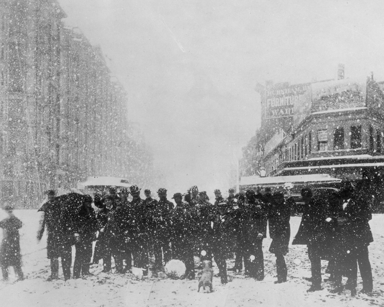 a black and white photo from December 31, 1881 showing a group of darkly-dressed people gathered together in the middle of Market Street.  The group is standing in a layer of white snow and the entire scene is filled with speckles of snowflakes.