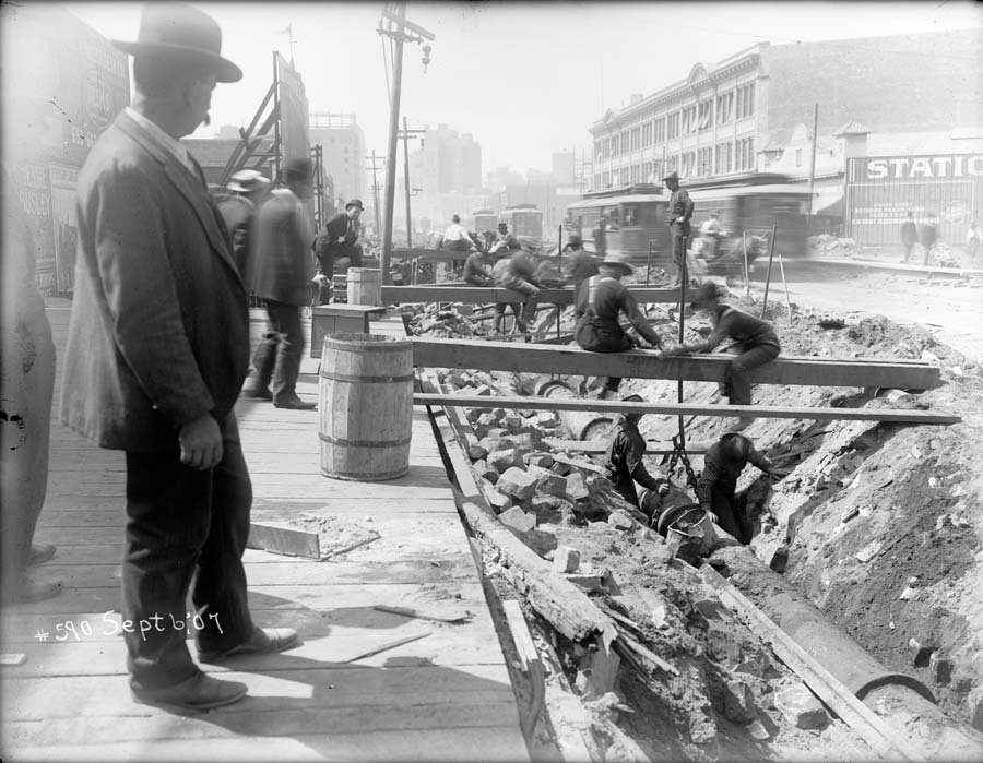 Black and white photograph taken on September 6, 1907 showing men in trench repairing water pipes.  The photographer is standing at the edge of a wooden plank sidewalk and there is a man with a large mustache and bowler hat standing near the camera at frame left. The men working in the trench are at lower center frame. Beyond and above them are streetcars passing on Market Street.