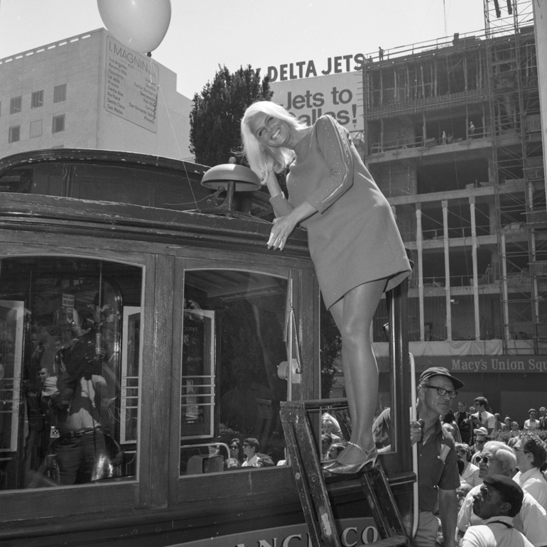 Black and white photograph of a woman standing on top of a ladder which is leaned against the front of a cable car in Union Square.  The woman is smiling as she leans towards the large bell on top of the cable car and cups her ear as if to hear the bell more closely. Taken on August 22, 1968 at the Cable Car Bell Ringing Contest.