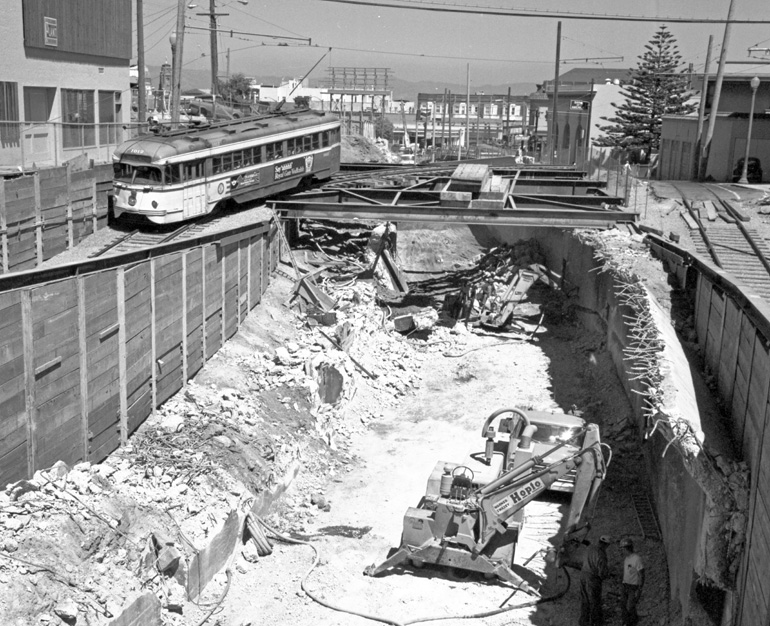 A streetcar passes over a large, open excavation on a set of raised tracks. This black and white image faces east on Market Street towards Castro Street, and was taken in 1973.