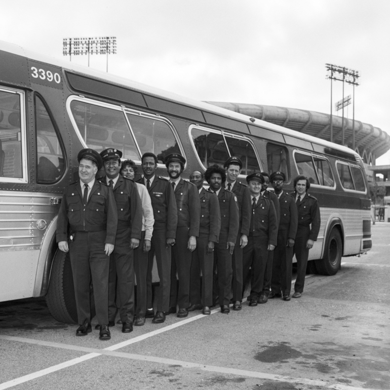 A black and white photo from the first Bus Roadeo in 1974 shows 12 Muni operators lined up standing in front of a 1960s-era bus. They're dressed in dark-colored Muni uniforms, including hats. The Candlestick Park stadium is in the background.
