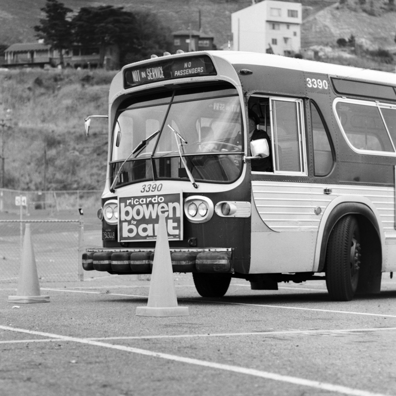 A black and white photo shows front-angled view of a Muni bus turning around cones in a parking lot during the first Muni Bus Roadeo in 1974.