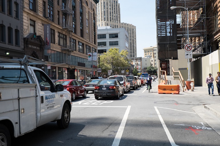 Trucks, cars and a bike travel north on a busy, one-way downtown street with construction on the right.