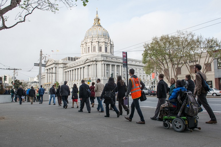 A group of people, one using a wheelchair, travels north on the west side of Van Ness Avenue with City Hall in the background.