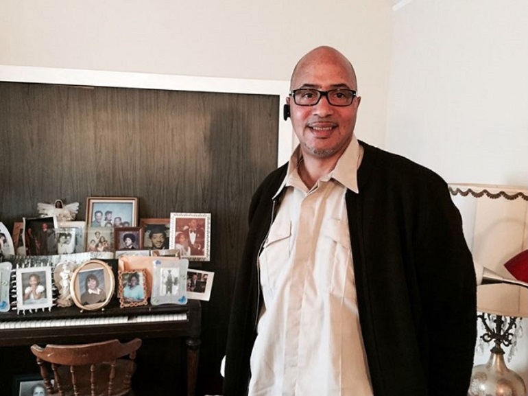 Matthew Smith standing in a living room in front of a piano with family photos on top.